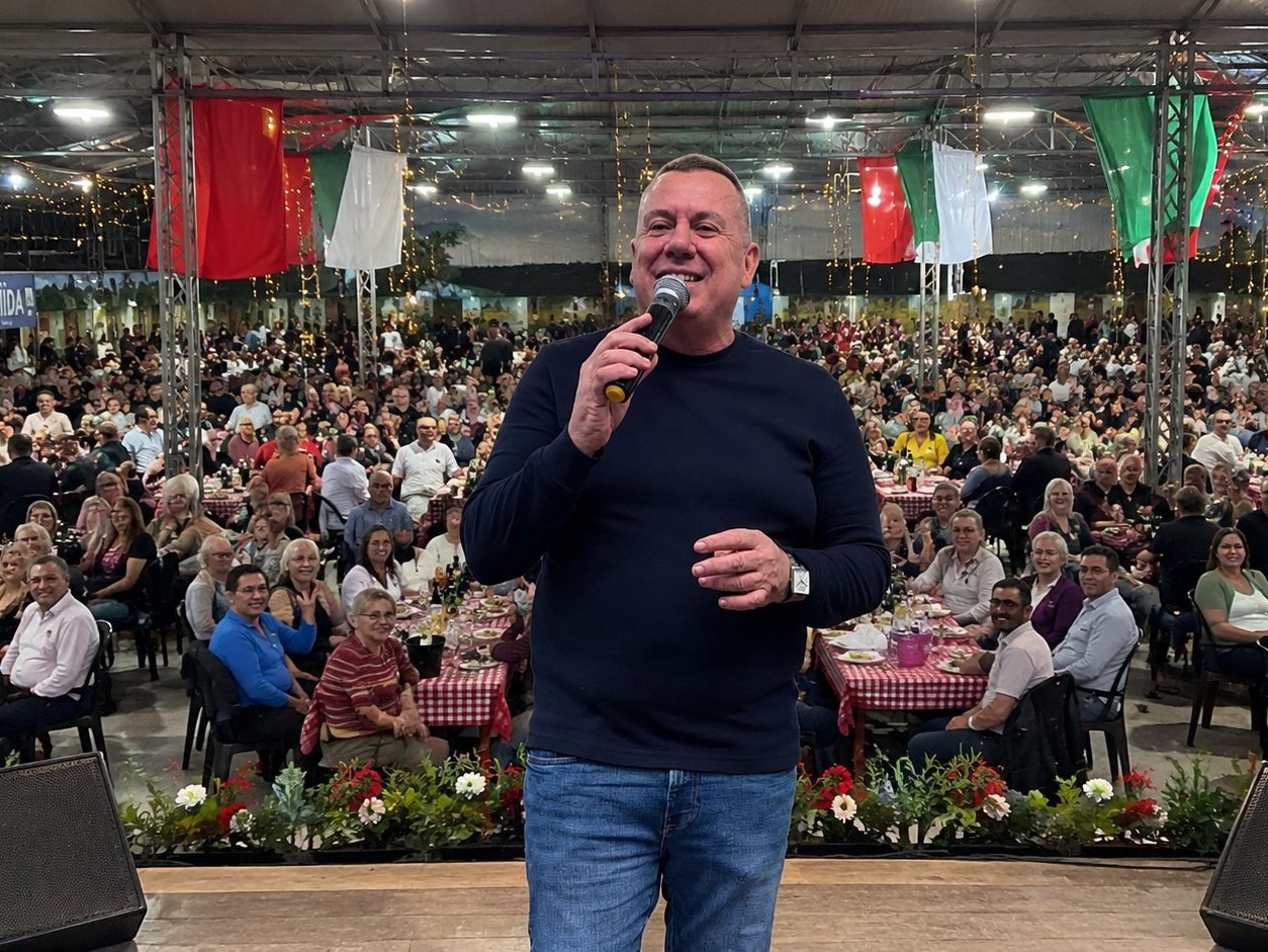 Público em uma grande festa típica, mesas cheias e bandeiras ao fundo